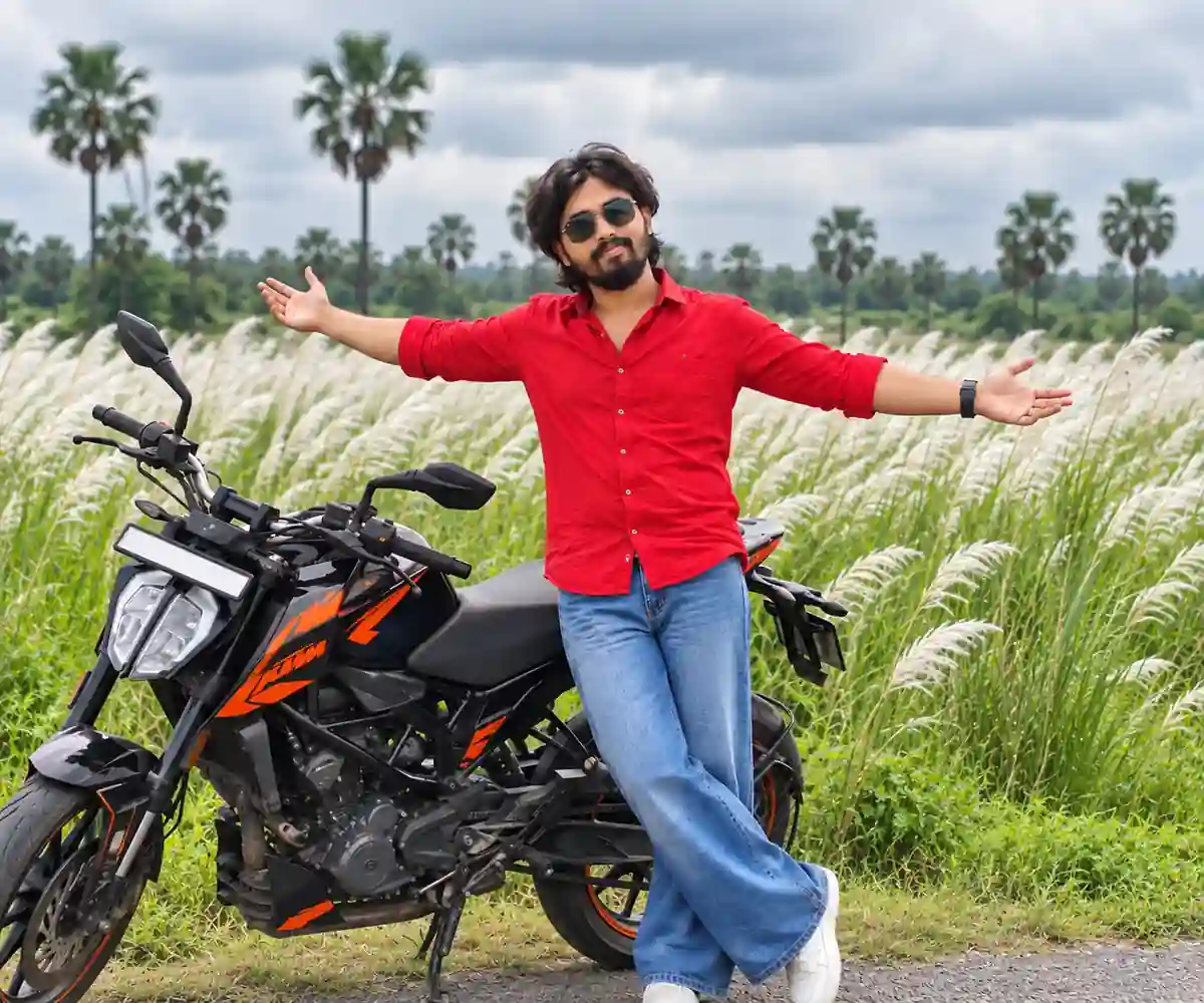Man in red shirt posing beside black and orange motorcycle in Kash flower field
